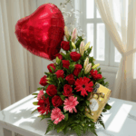 Arreglo floral con rosas rojas, gerberas rojas y rosas, lirios claros, globo metálico rojo en forma de corazón y caja de chocolates sobre mesa blanca frente a ventana con cortinas claras