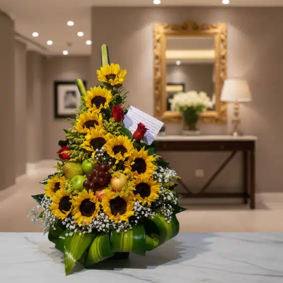 Arreglo floral dorado con girasoles, rosas rojas, uvas, manzanas y peras verdes, sobre una mesa de mármol en un elegante reci