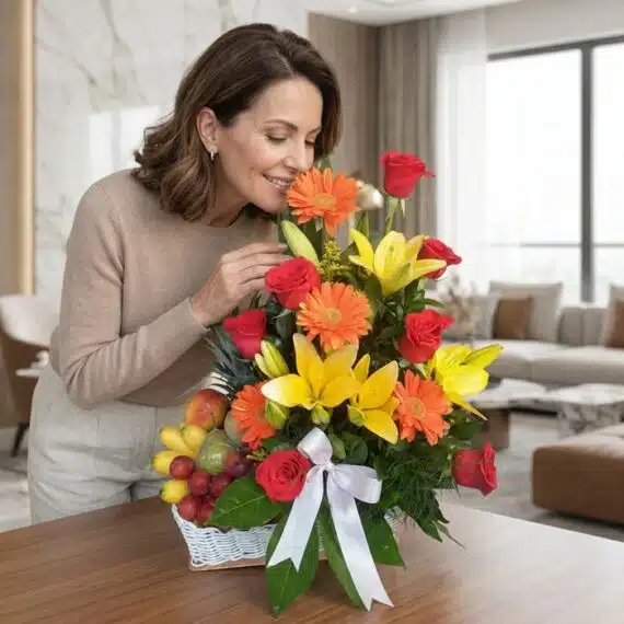 Mujer elegante de mediana edad admirando y oliendo un Arreglo Floral con Frutas Quitana, con rosas rojas, lirios amarillos y