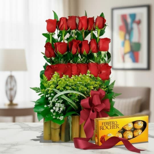 Arreglo Floral Musca con rosas rojas vibrantes, follaje verde, base de bambú y caja de chocolates Ferrero Rocher sobre mármol