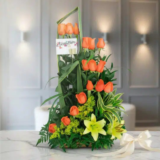 Arreglo floral Romeo con rosas naranjas, lirios amarillos y follaje verde sobre mármol, listo para regalar.