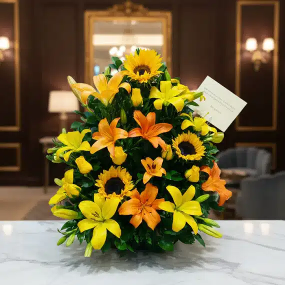 Arreglo Floral Tropical con Girasoles y Lirios para Decoración Elegante Arreglo floral tropical vibrante con girasoles, lirios amarillos y naranjas, y follaje verde, sobre una superficie de mármol