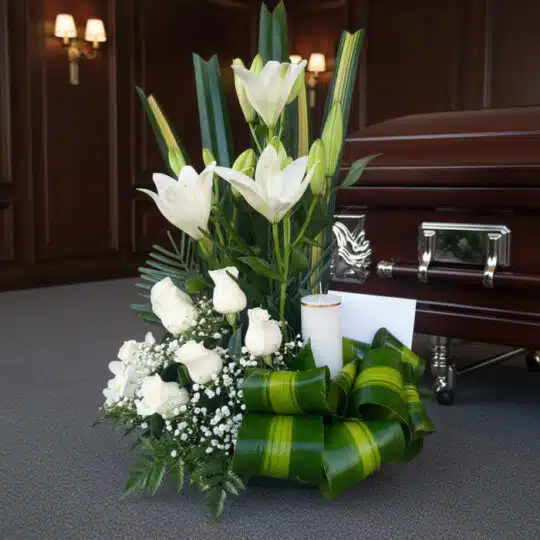 Arreglo floral fúnebre Claridad con lilas y rosas blancas, follaje verde, vela y tarjeta, junto a un ataúd en sala de velació