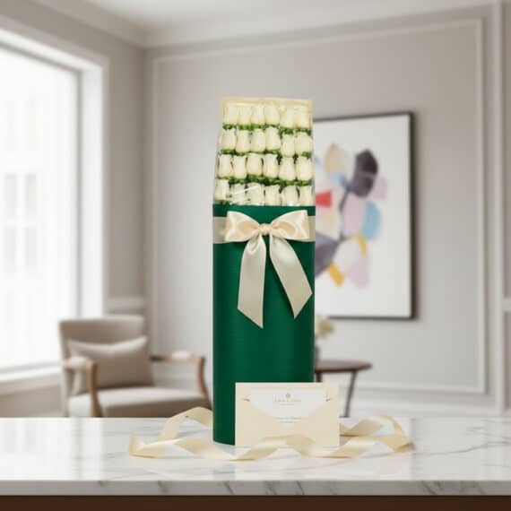 Caja cilíndrica verde con 24 rosas blancas, lazo satinado color crema, tarjeta de regalo y cinta sobre mármol.