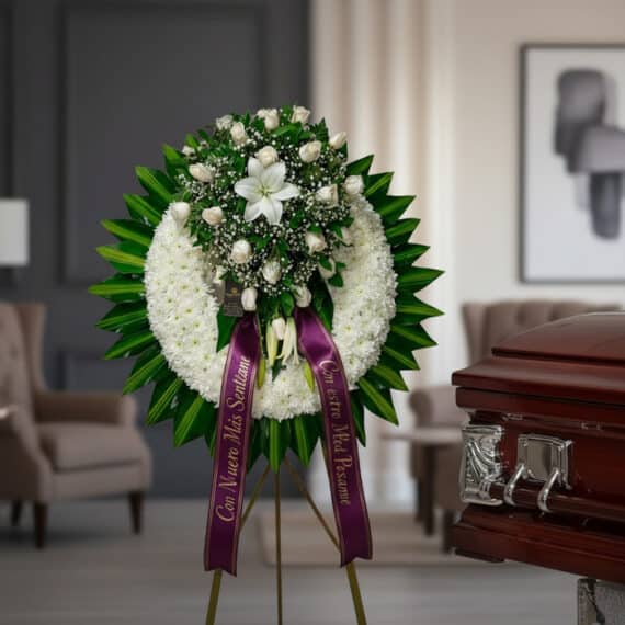 Impresionante corona fúnebre con rosas blancas, lirios y follaje verde, sobre un caballete, con cinta morada de condolencia,