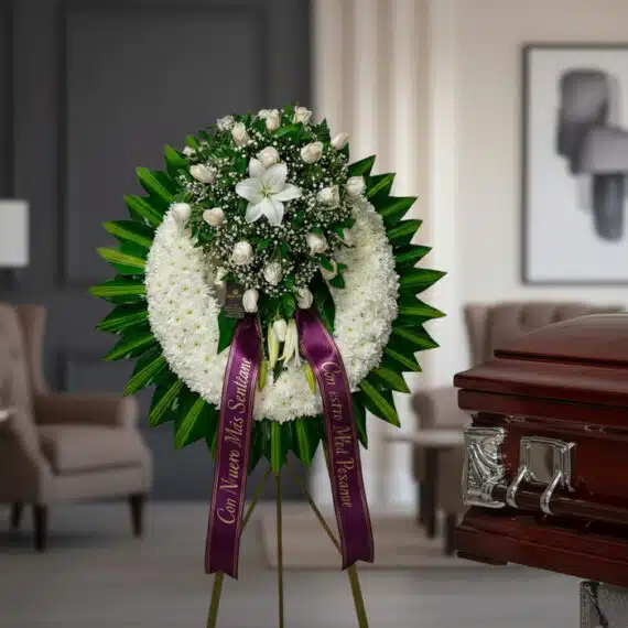 Impresionante corona fúnebre con rosas blancas, lirios y follaje verde, sobre un caballete, con cinta morada de condolencia,