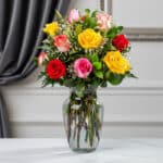 Jarrón de cristal transparente con un ramo de 12 rosas de colores variados: rojas, amarillas, rosas y blancas, con follaje ve