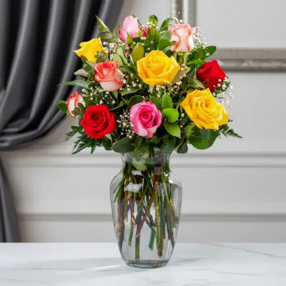 Jarrón de cristal transparente con un ramo de 12 rosas de colores variados: rojas, amarillas, rosas y blancas, con follaje ve