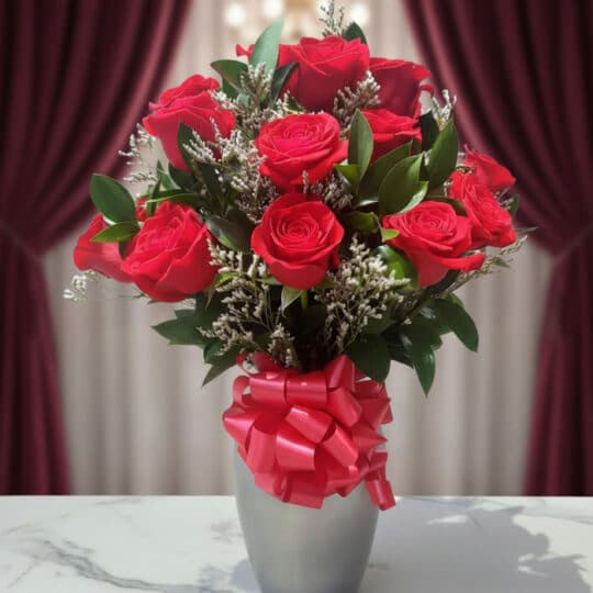 Jarrón plateado con doce rosas rojas frescas y follaje verde, atado con un lazo rojo brillante, sobre una mesa de mármol blan