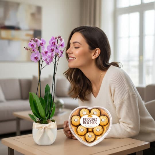 Mujer sonriendo y oliendo una orquídea GALA morada en maceta blanca con lazo, junto a una caja de chocolates Ferrero Rocher e