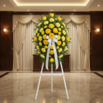 Pedestal fúnebre Legado Sereno con rosas y claveles amarillos, lazo blanco, en sala de velación elegante con cortinas y luz c