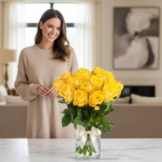 Mujer sonriente admirando un arreglo floral de 18 rosas amarillas en un jarrón de cristal con lazo, en un hogar de diseño luj